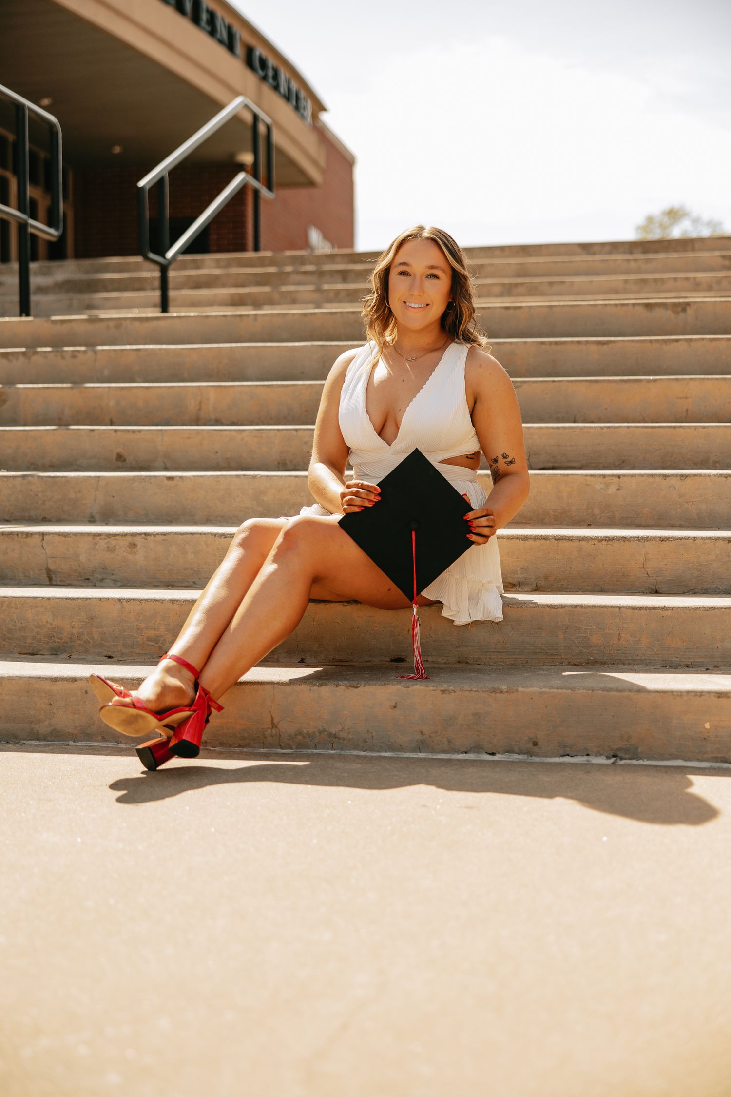 Graduate in white dress poses casually on concrete steps with red shoes.