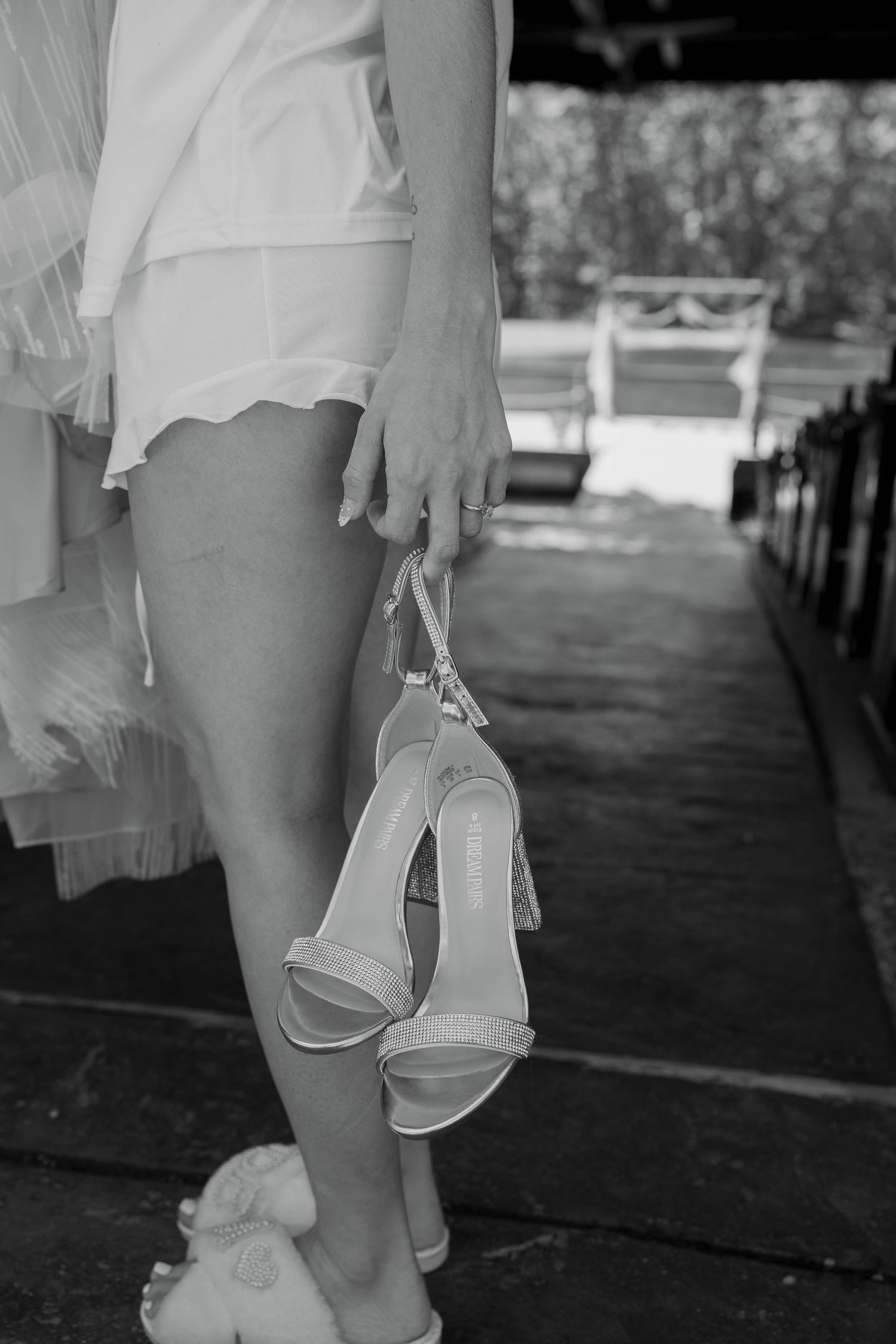 Bride holding white heeled sandals in hand, wearing short wedding dress, black and white photo at outdoor venue.
