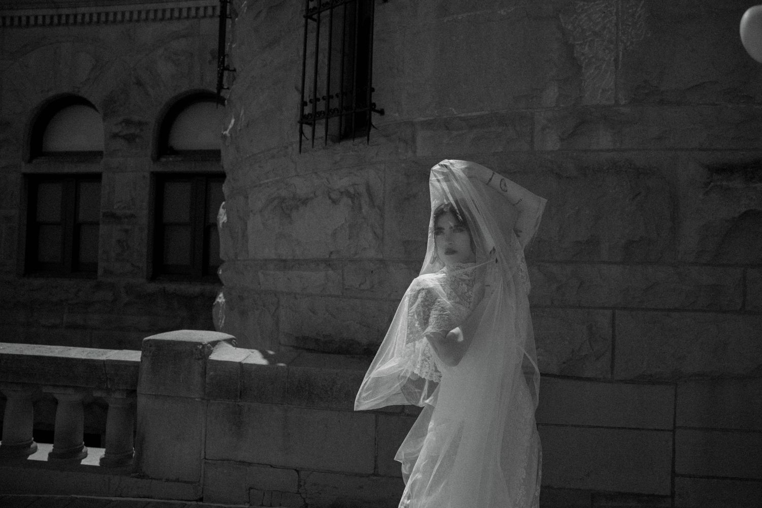 Black and white photo of veiled bride standing beside stone building exterior.