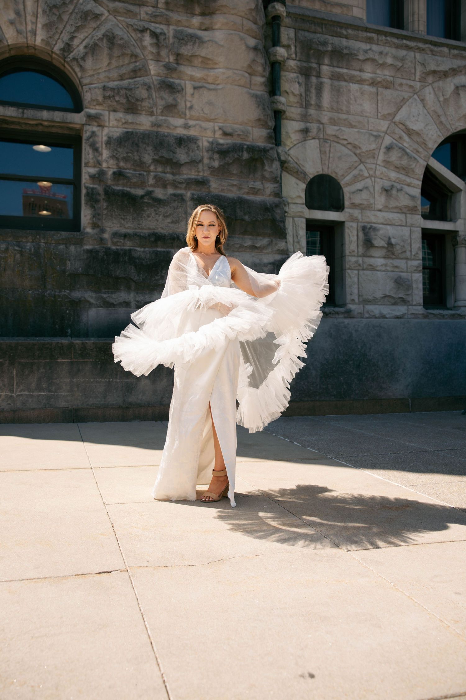 Bride twirls dramatic ruffled tulle veil in sunlight wearing fitted white gown outside historic stone building.