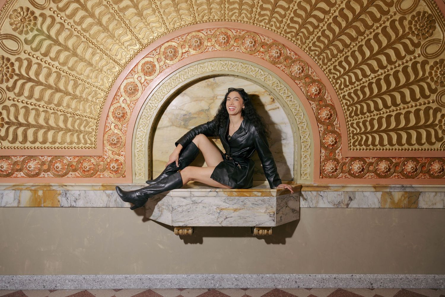 Woman in black leather outfit and boots poses in ornate arched niche with golden decorative molding.