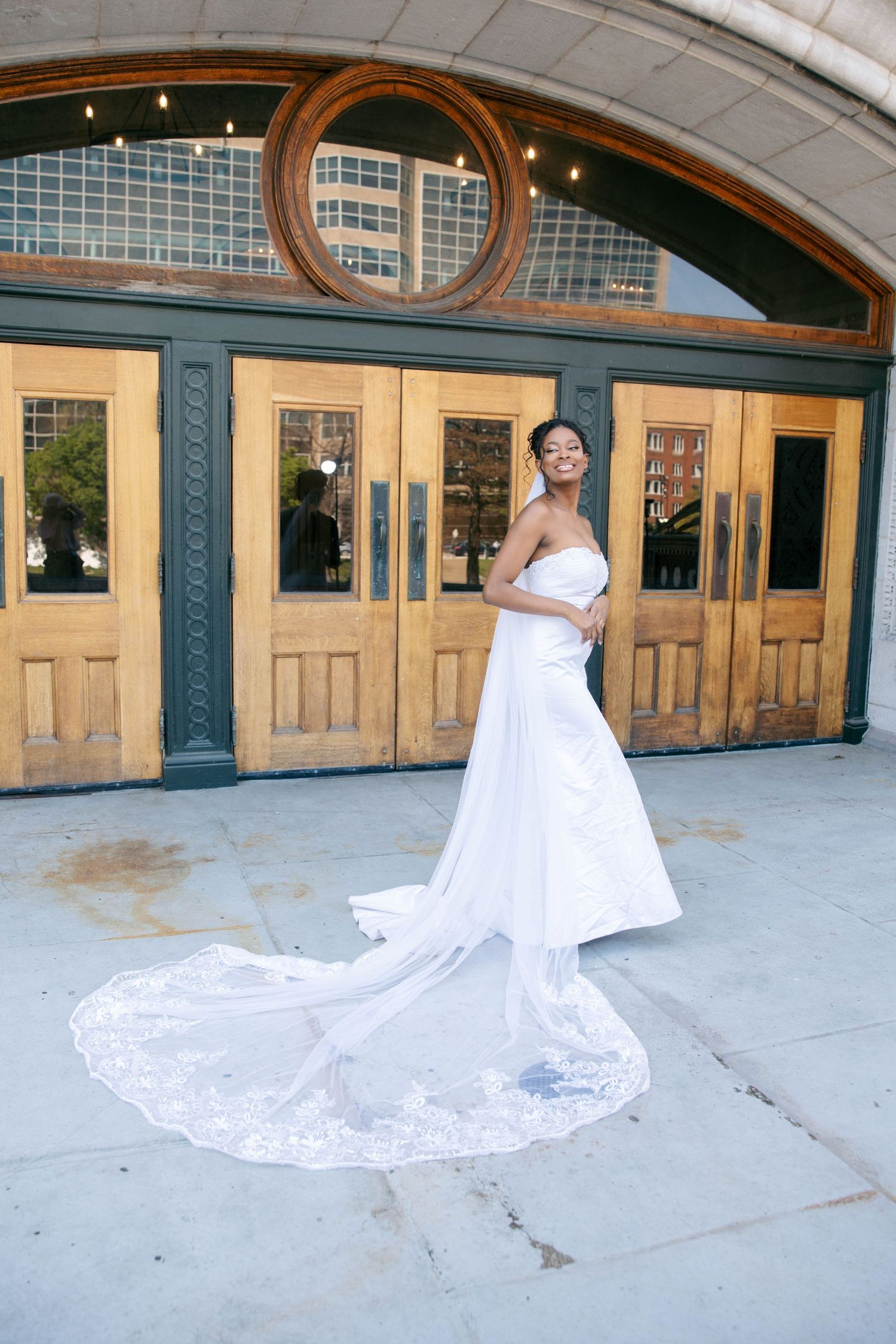 Elegant bride in a strapless white gown with lace-trimmed cathedral train poses before ornate wooden doors.