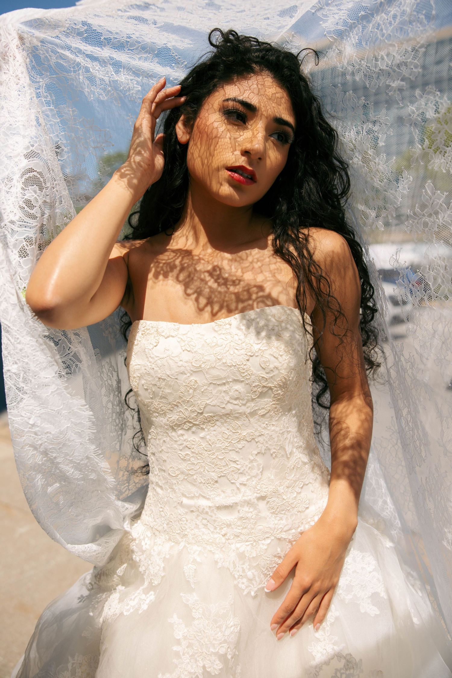 Bride in white lace gown under sheer veil with sunlight casting floral shadows on her skin outdoors.