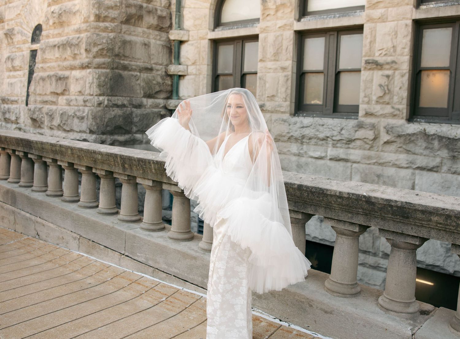 Bride in white lace gown and feathered veil poses gracefully outside a historic stone building.