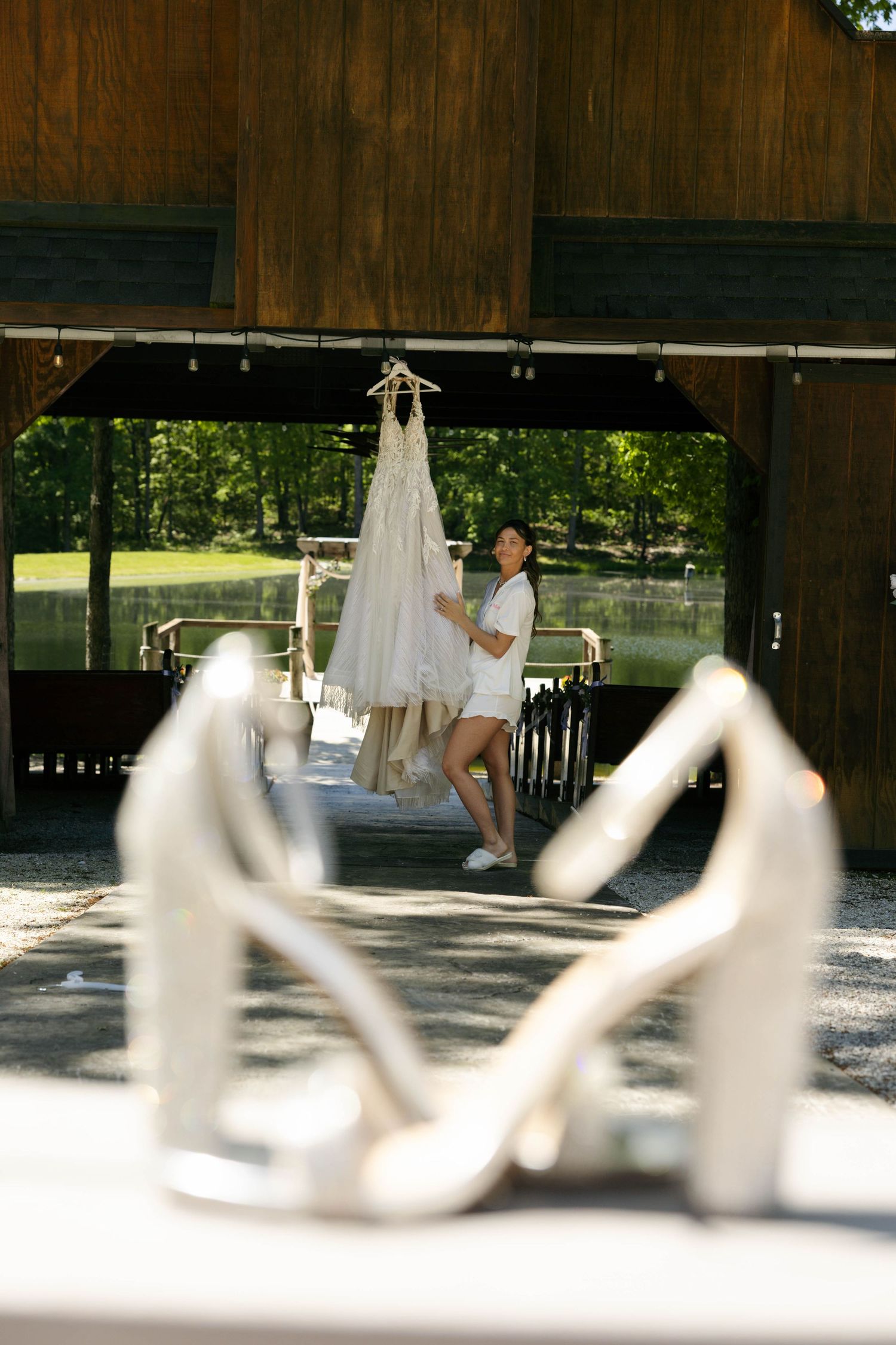 Bride admiring her wedding dress hung in a barn, with elegant white heels in the foreground.