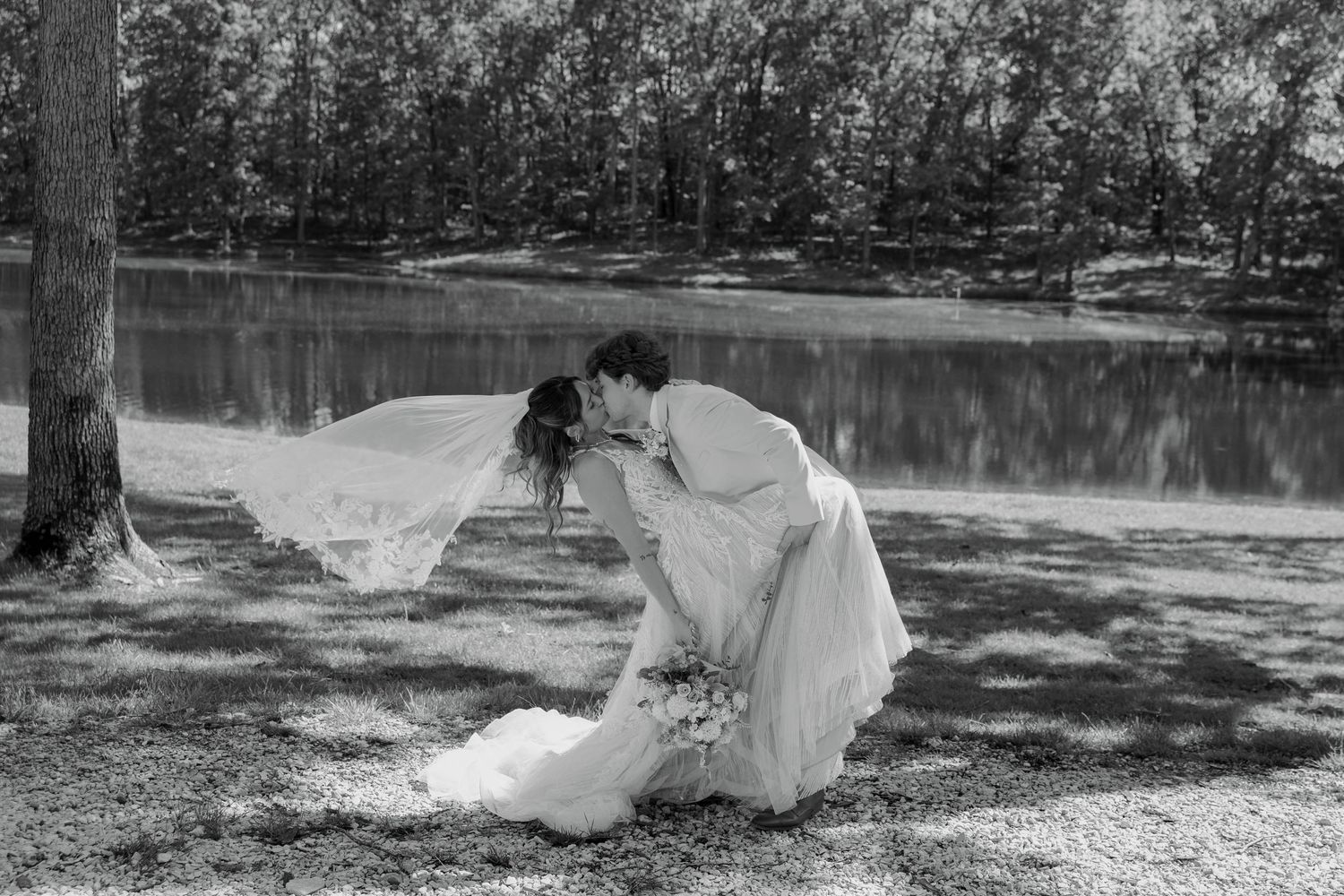 Black and white photo of a couple sharing a romantic dip kiss in wedding attire near a serene lake surrounded by trees.