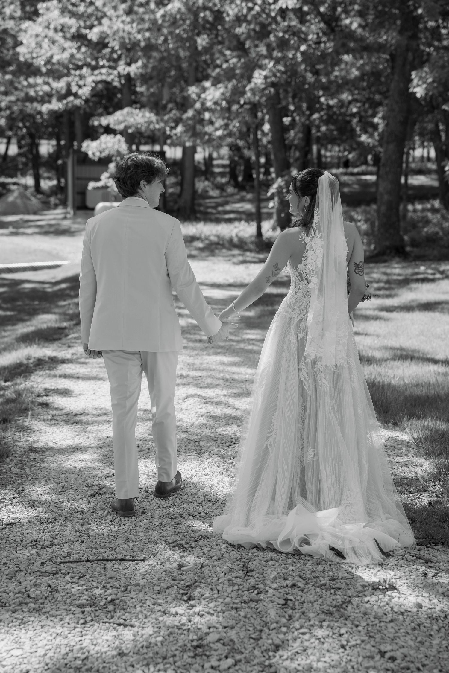 Black and white photo of a bride and groom holding hands walking through a tree-lined path on their wedding day.