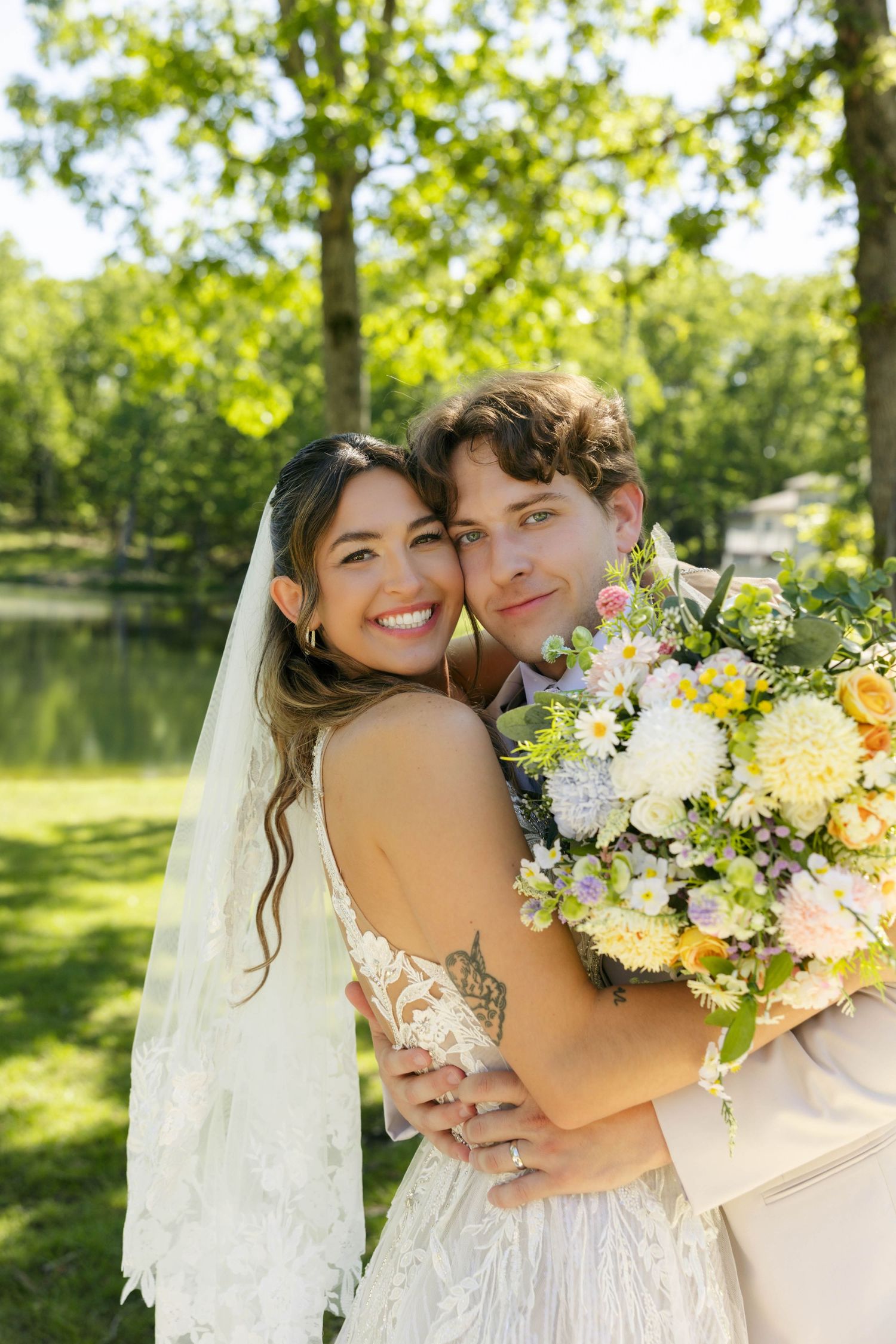 Smiling bride in lace dress and veil embraces groom outdoors, holding yellow and white floral bouquet in a sunny park.