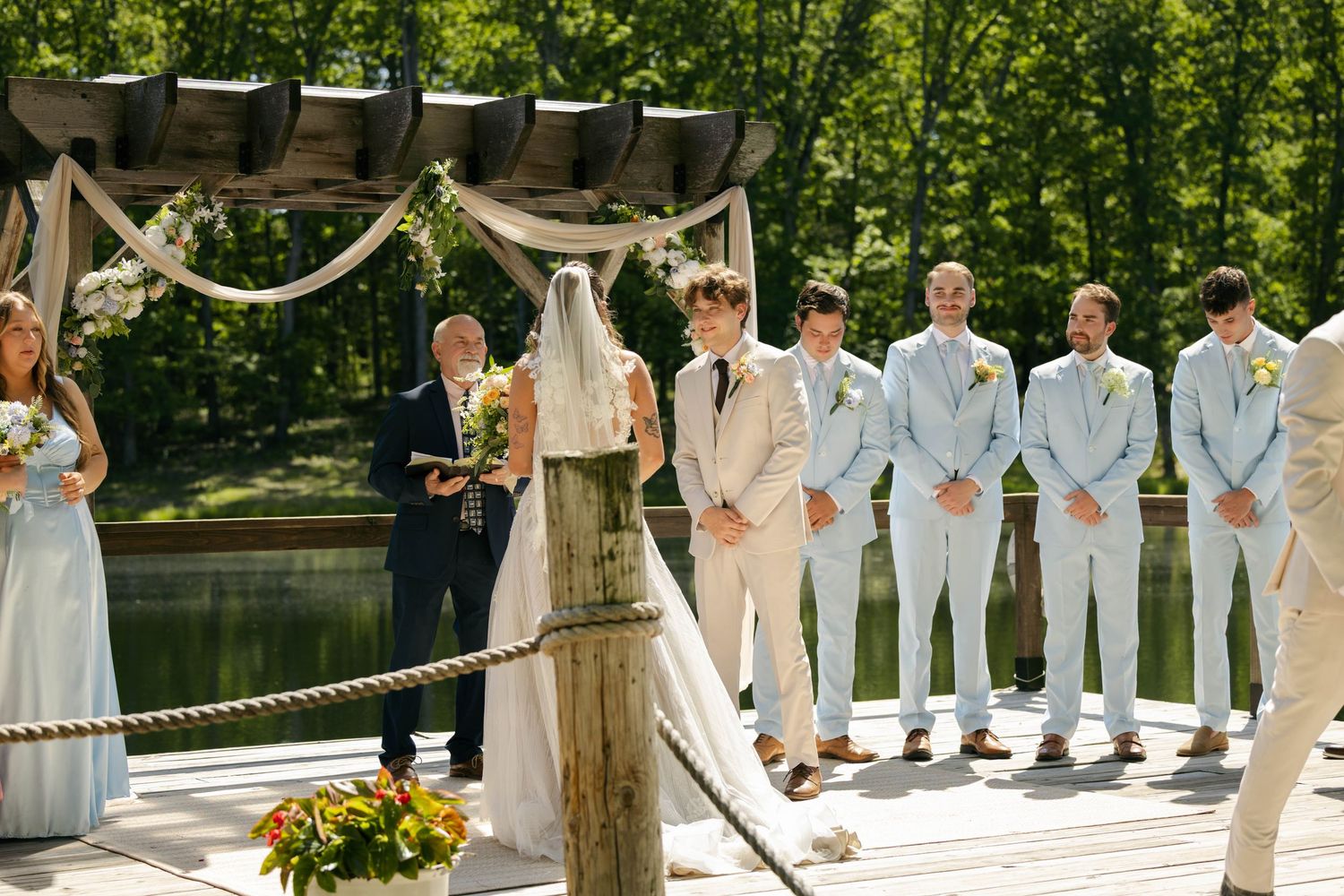 Bride and groom exchange vows at outdoor dock wedding with groomsmen in cream and blue suits beside lake.