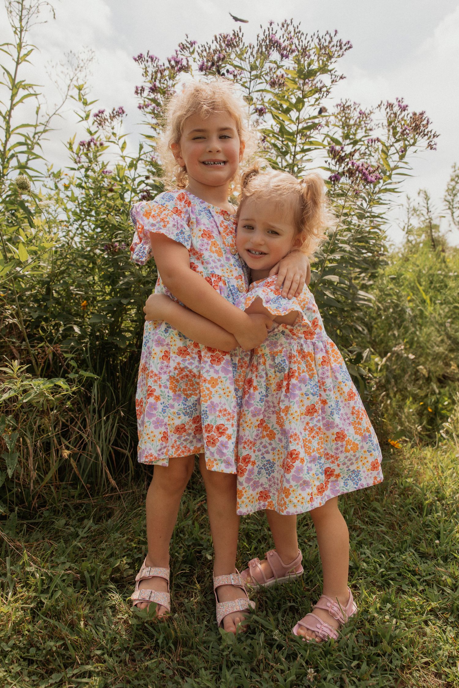 Two matching floral dresses twirl together in a garden setting.