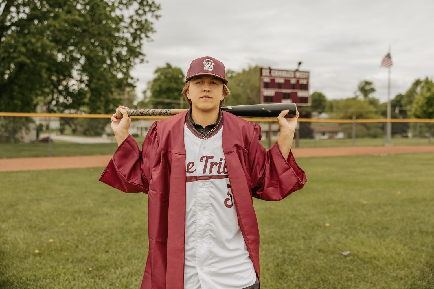 Graduate in maroon gown stands on baseball field under cloudy sky.