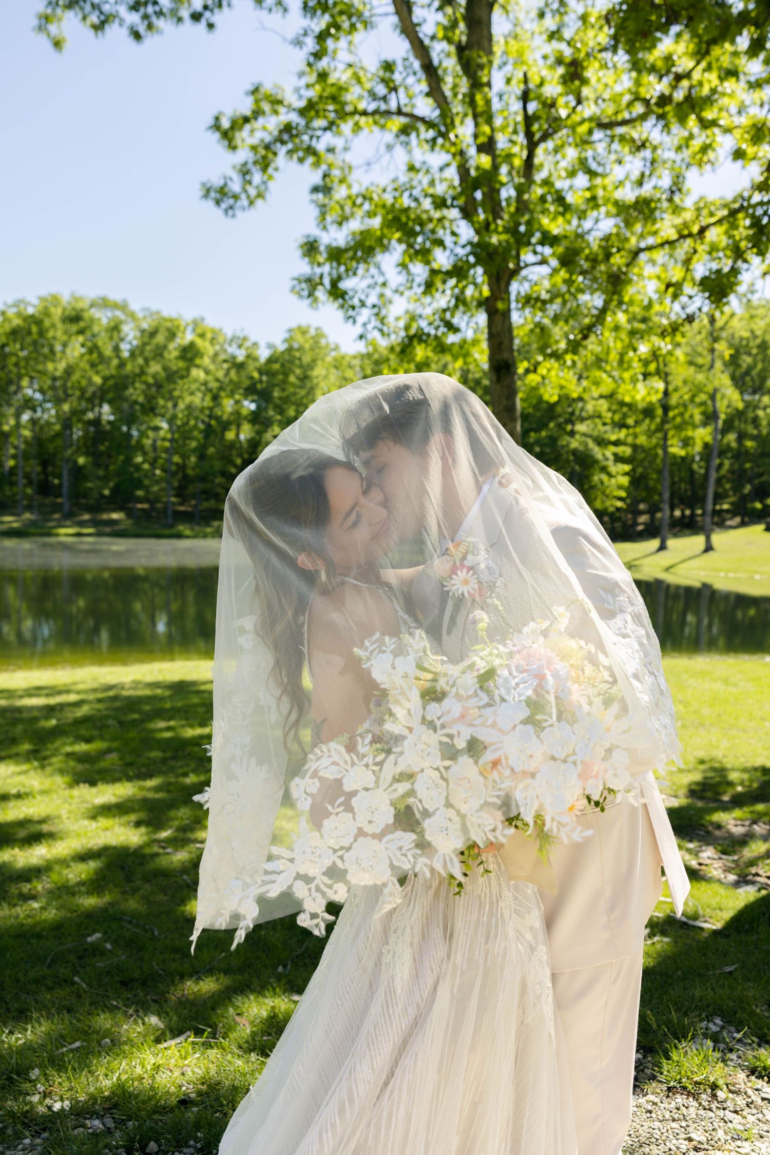 Bride and groom share a kiss under a lace veil outdoors, holding a white floral bouquet near a pond.