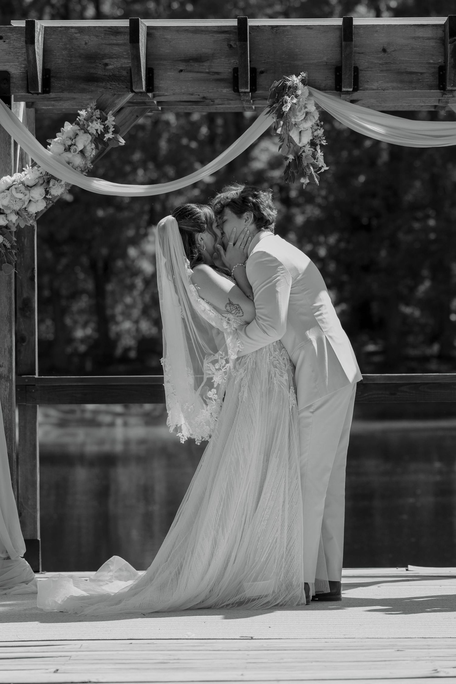 Black and white photo of a bride and groom sharing their first kiss under a floral arch by a lake.