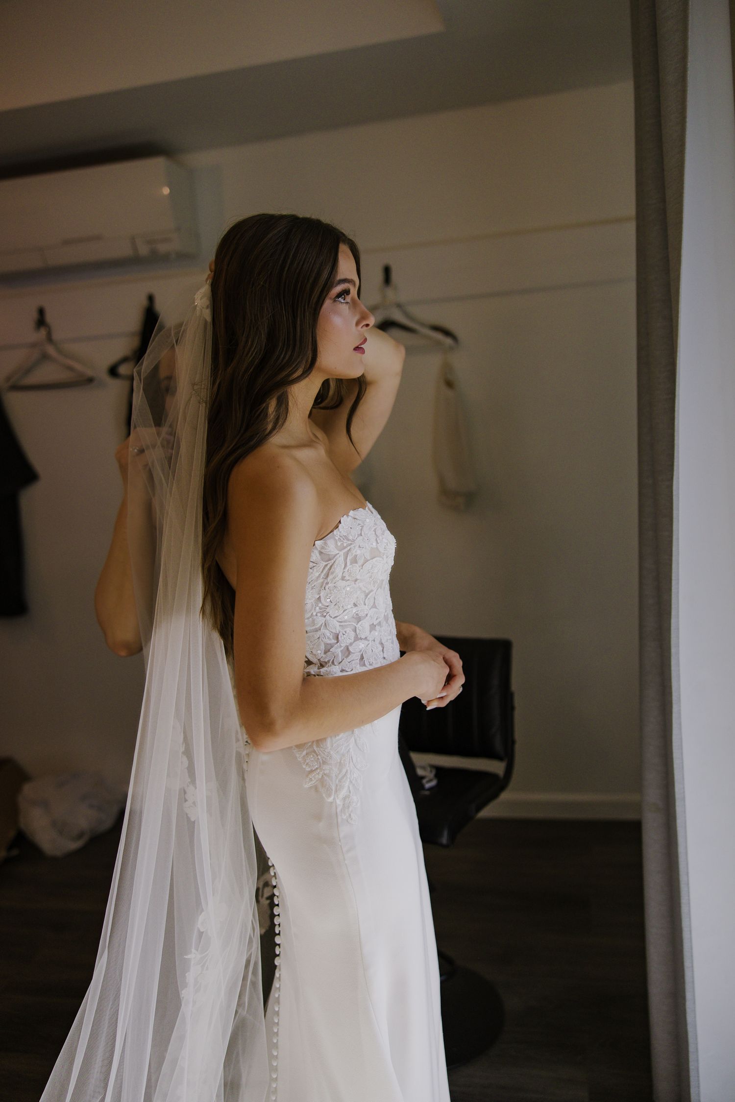 A bride in a lace wedding dress and veil stands in a dressing room.
