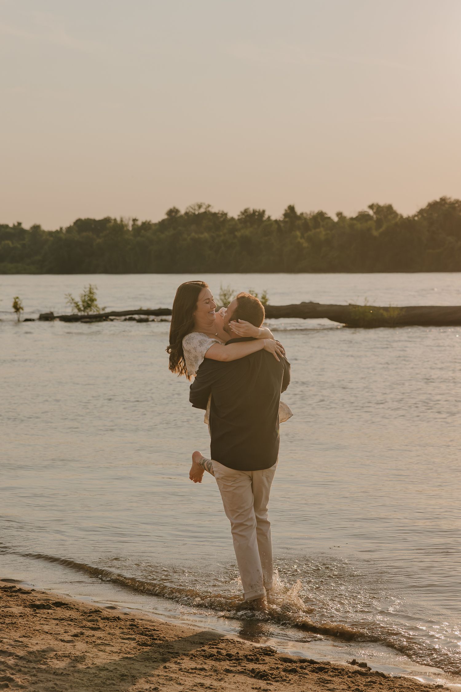 Couple embraces on a sandy beach at sunset with calm waters and trees in the background.