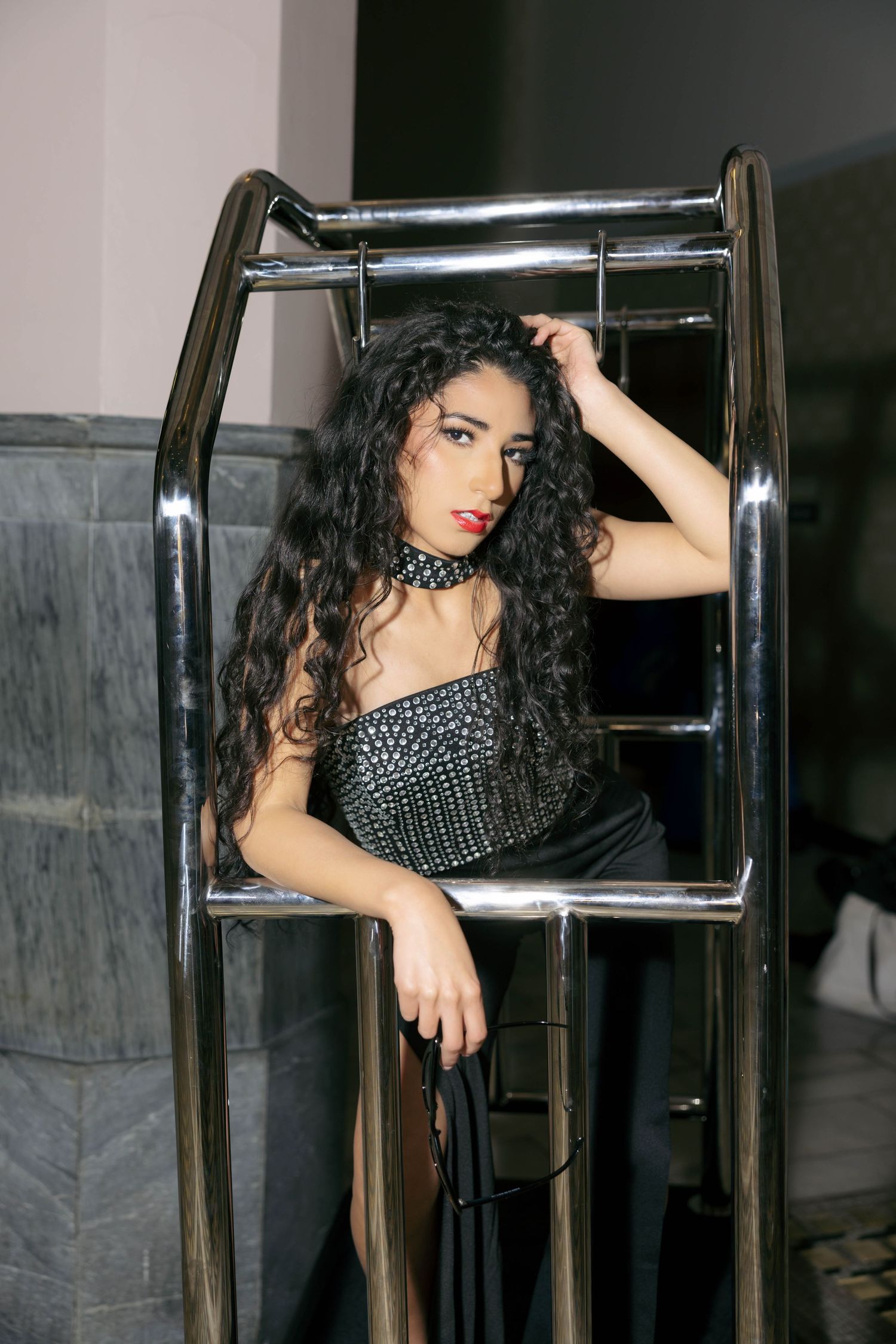 Young woman with long curly black hair poses in a studded black corset top and choker between chrome metal railings.