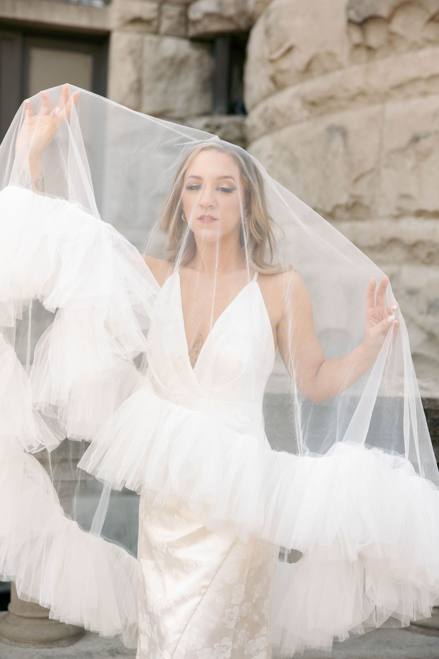 Bride in white tulle wedding dress holds flowing veil aloft outdoors near stone architecture.