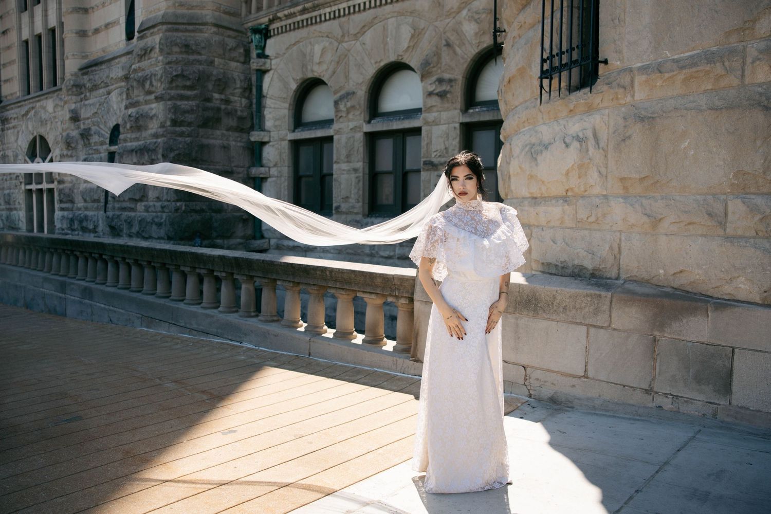 Bride in sleek white gown with flowing veil poses on sunlit terrace of a grand historic building.