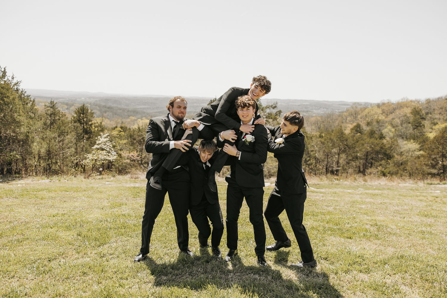 Groomsmen in black suits playfully lift someone during outdoor wedding photos.