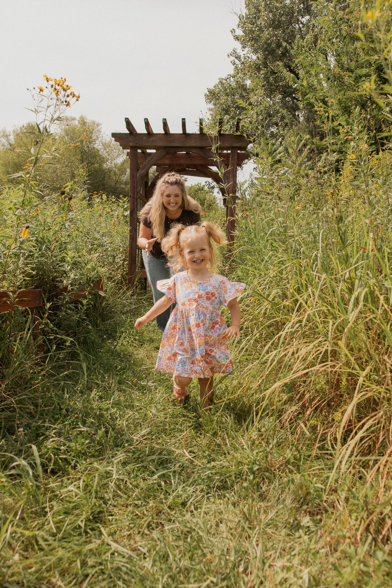A floral dress-clad figure runs through tall grass near a wooden pergola.