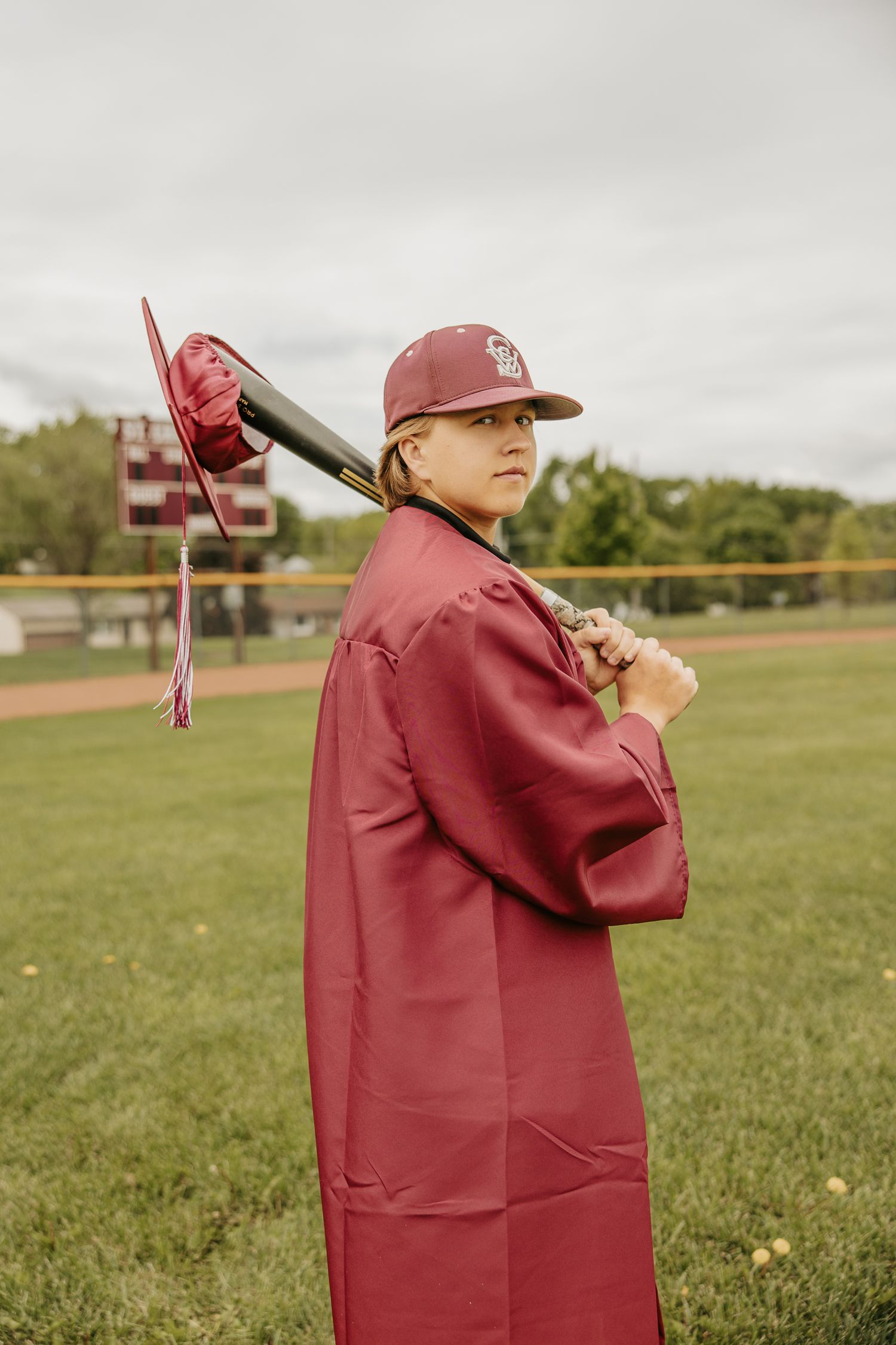 Person in maroon graduation gown holds diploma case in outdoor field setting.