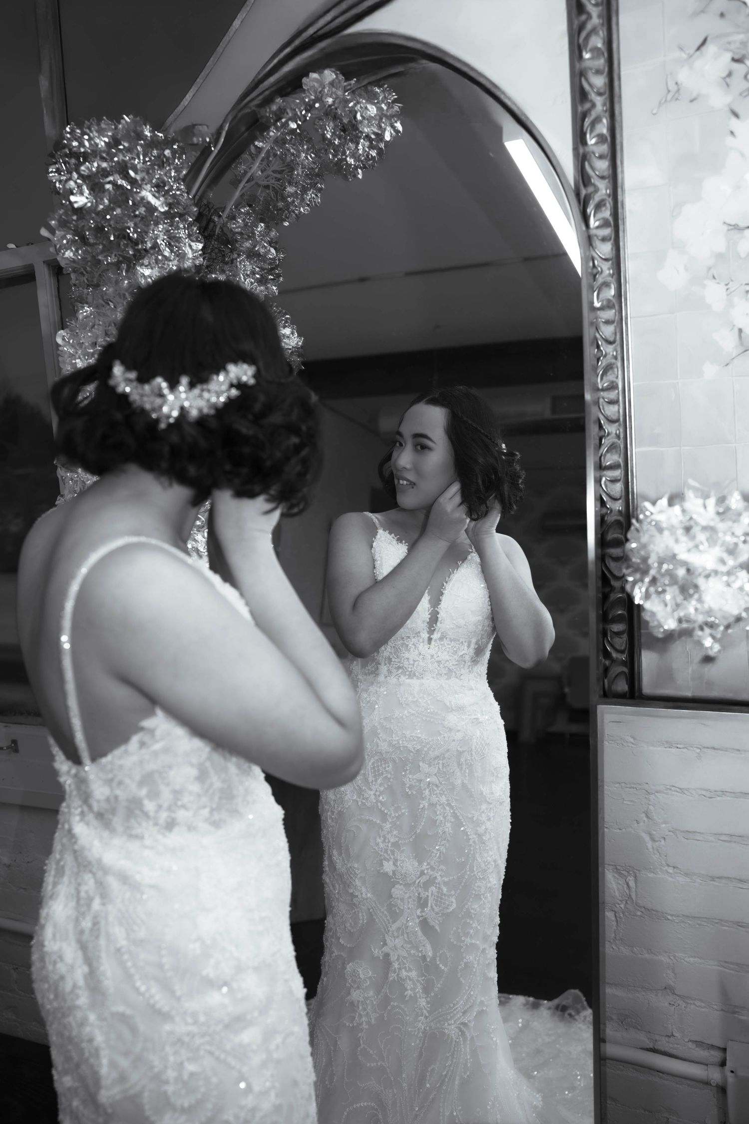 Bride in lace wedding gown adjusts earrings while gazing at her reflection in an ornate mirror, black and white photo.