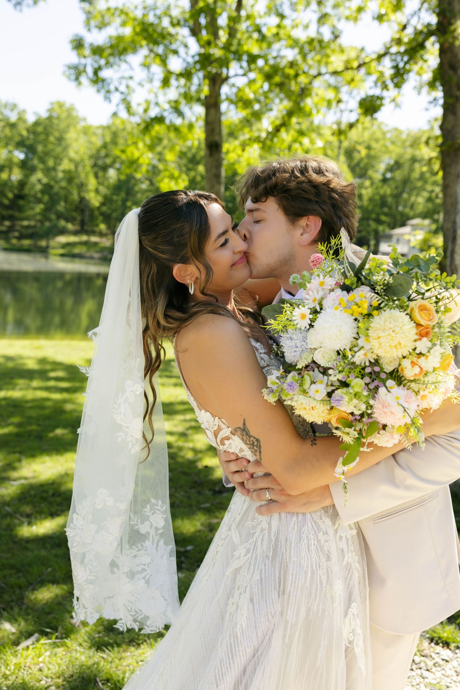 Bride and groom sharing a kiss outdoors, holding a pastel floral bouquet, with lush green trees and a pond in the background.