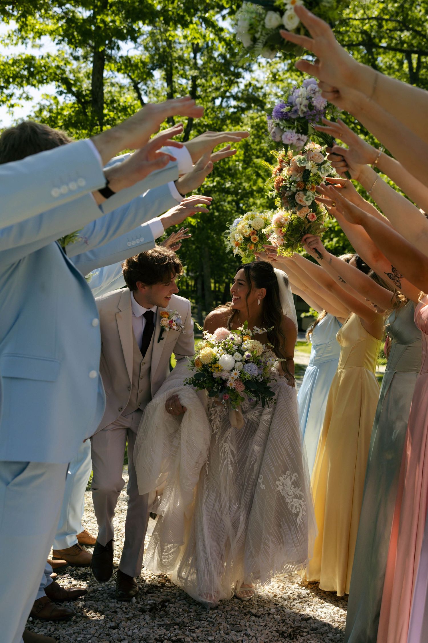 Bride and groom walk through an arch of hands held by wedding guests outdoors among lush green trees.