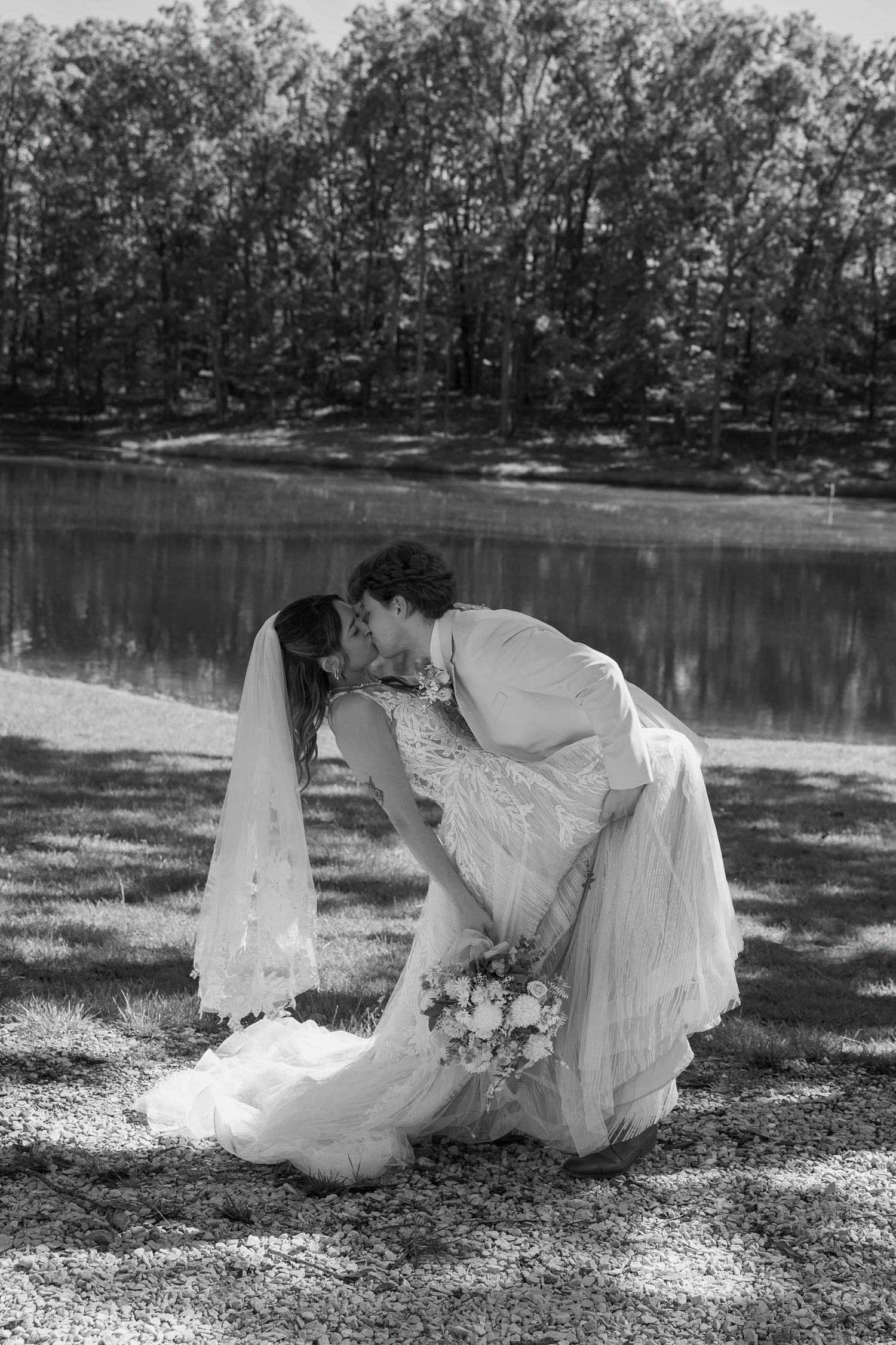 Black and white photo of a bride and groom sharing a romantic dip kiss outdoors near a lake.