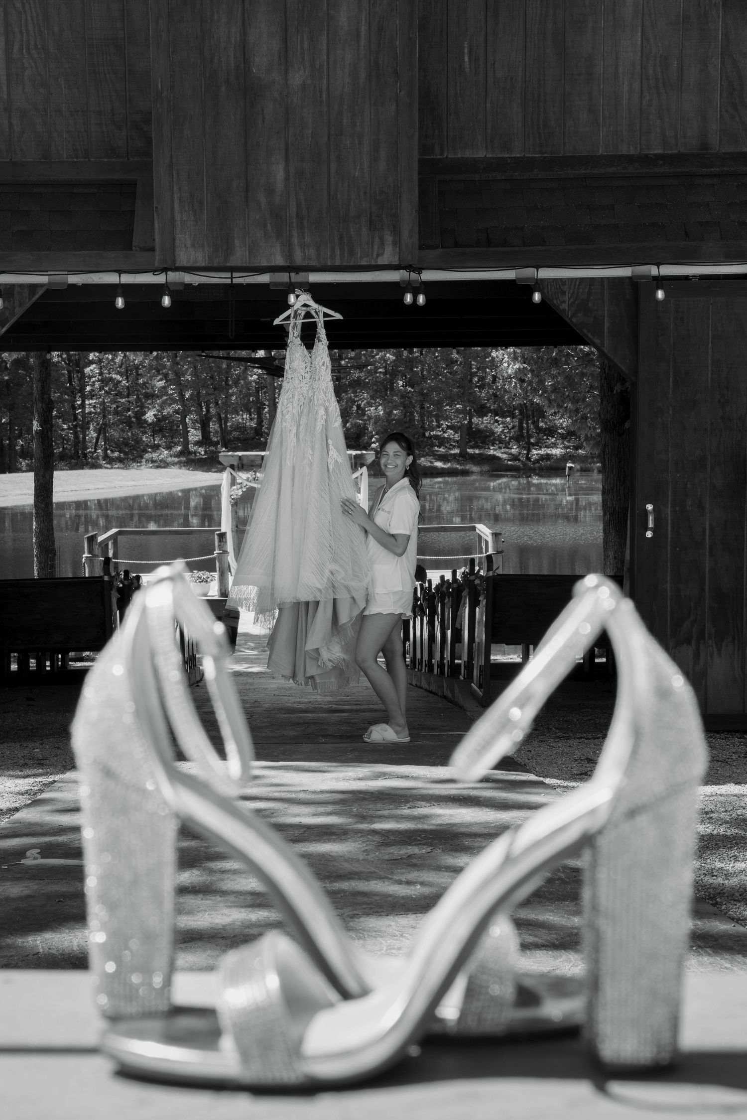Black and white photo of bride holding wedding dress with sparkly heels in foreground at outdoor barn venue.