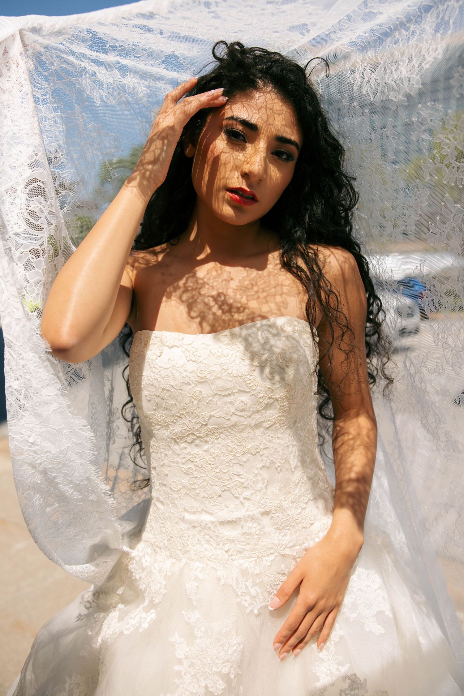 Bride in strapless white lace gown under sheer veil with dappled sunlight on face.