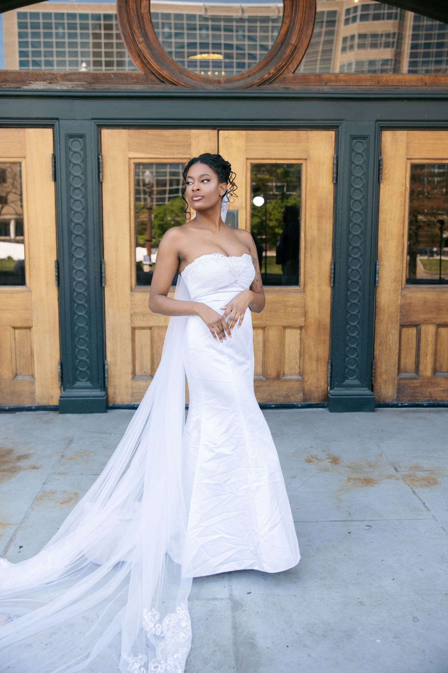 Elegant bride in strapless white gown with flowing train poses before wooden doors in urban setting.