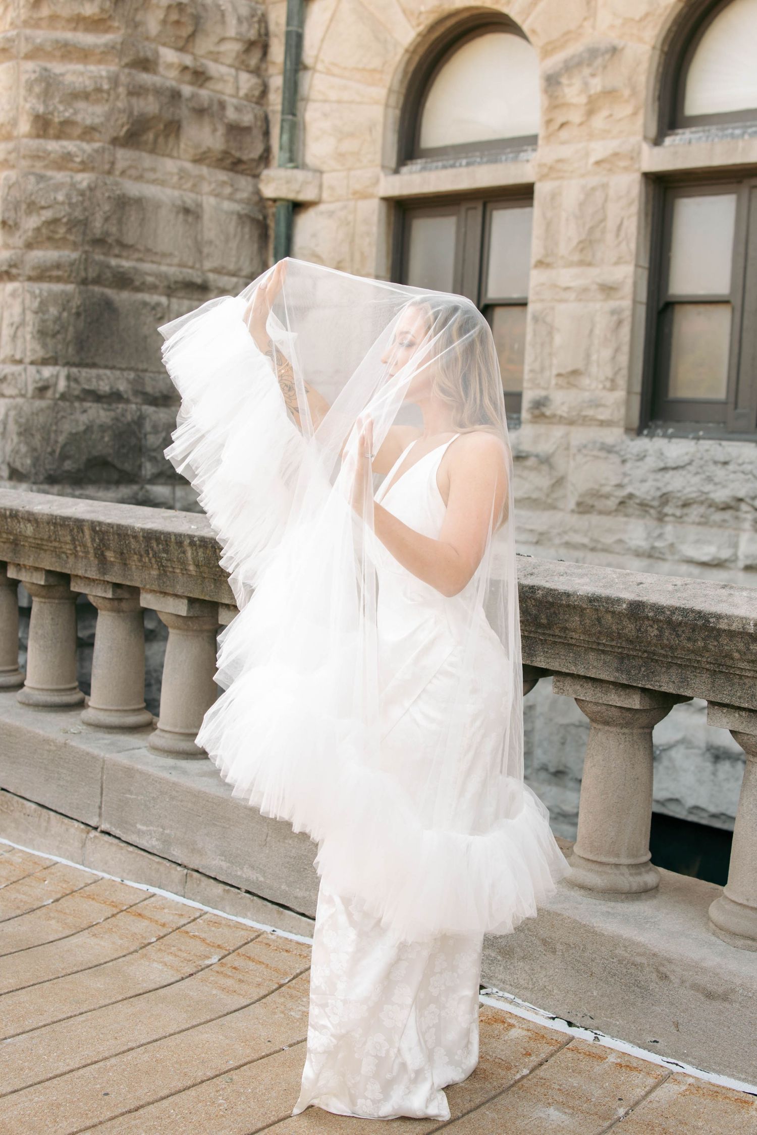 Bride in lace wedding dress and flowing veil poses on stone balcony of historic building.