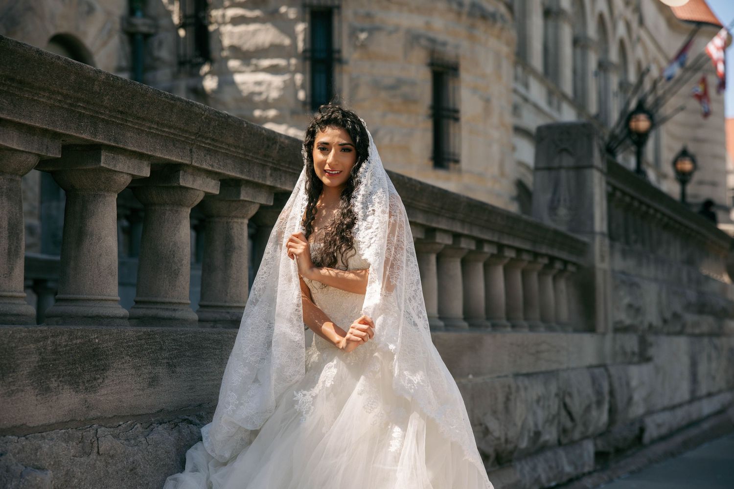 Bride in elegant white lace wedding gown and veil poses on a sunlit European street.