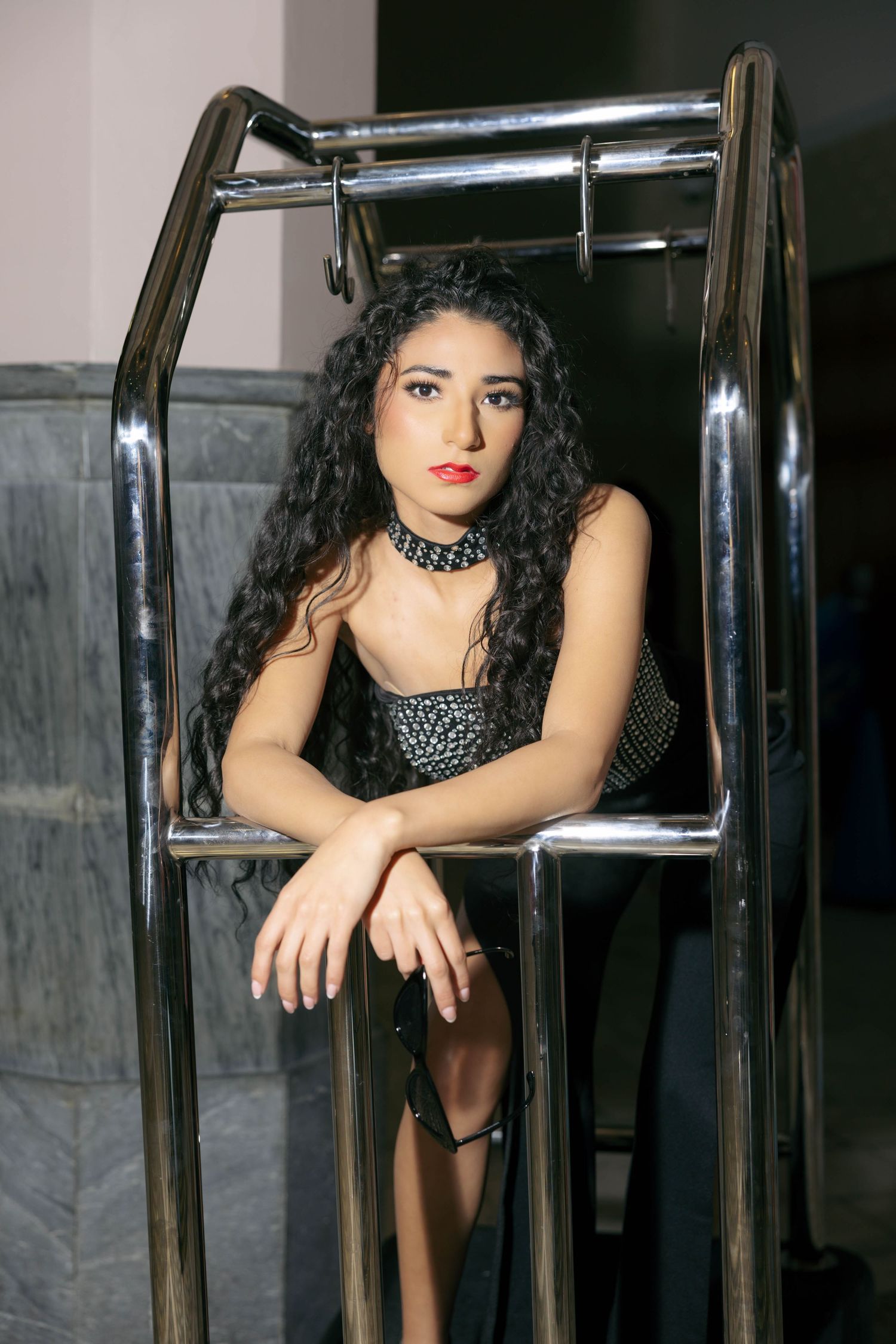 Young woman with curly black hair in a black outfit and choker poses inside a metal frame structure.