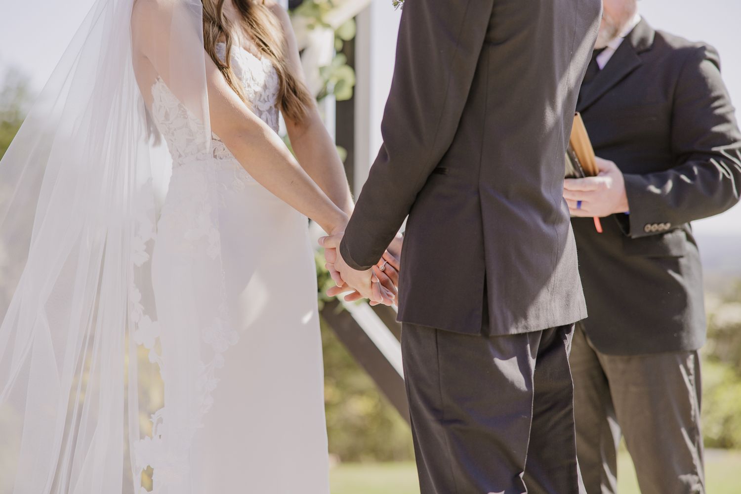 Bride and groom hold hands during sunny outdoor wedding ceremony.