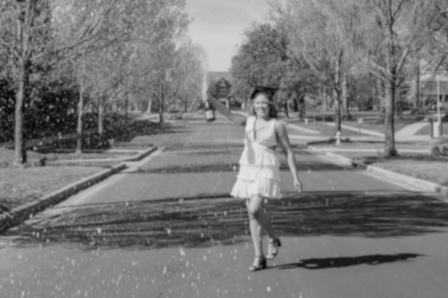 Black and white vintage style photo of someone twirling in a dress on a tree-lined street.