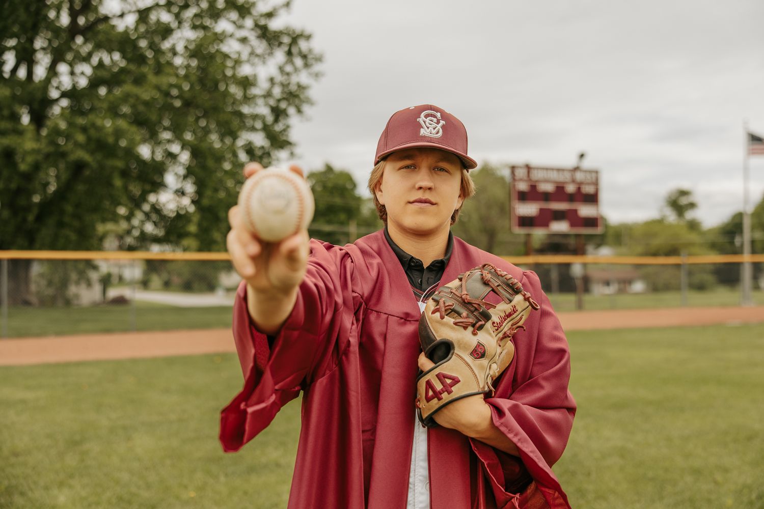 Graduate in maroon cap and gown poses with diploma on sports field.