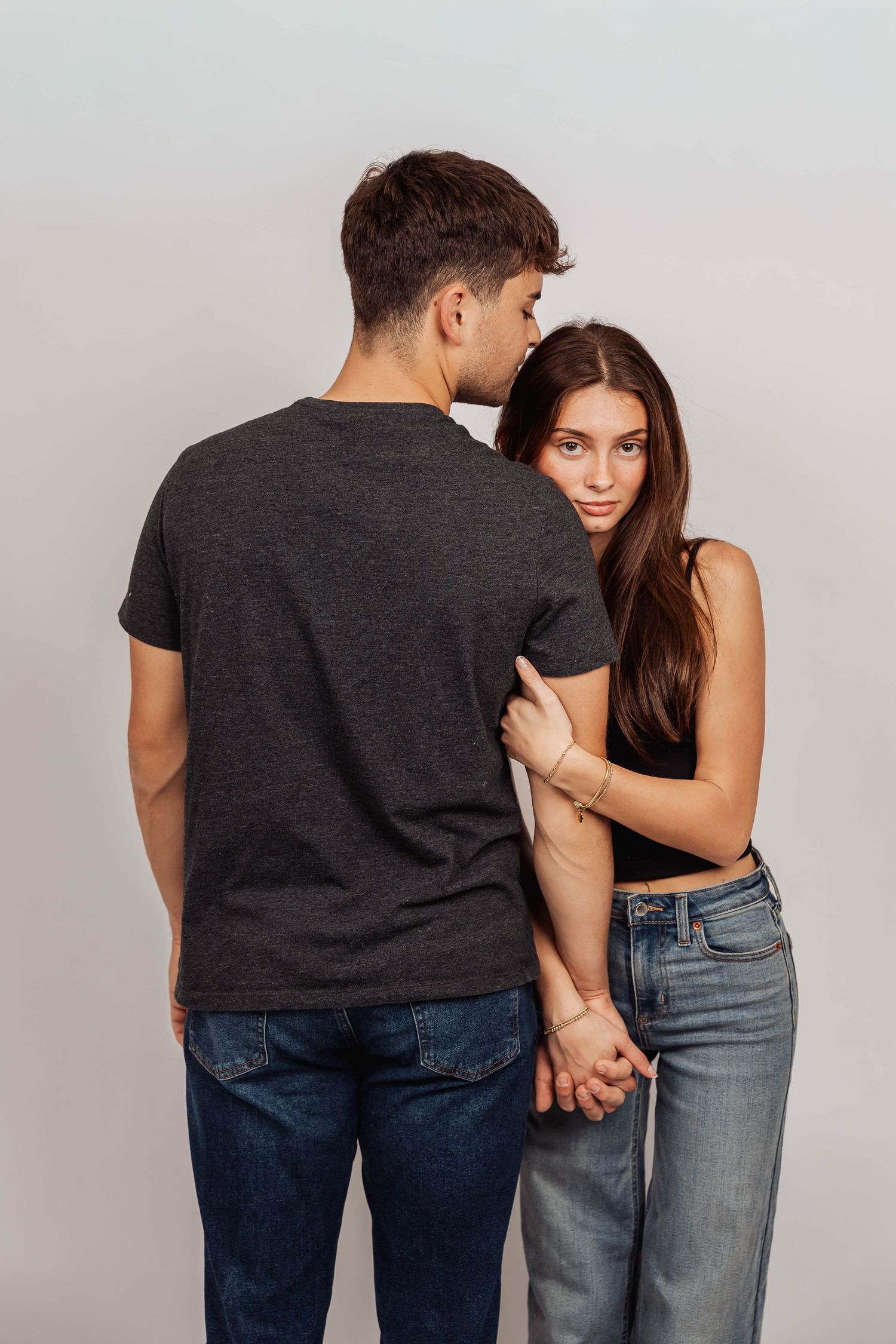 Two people in jeans and dark tops pose intimately against white background.