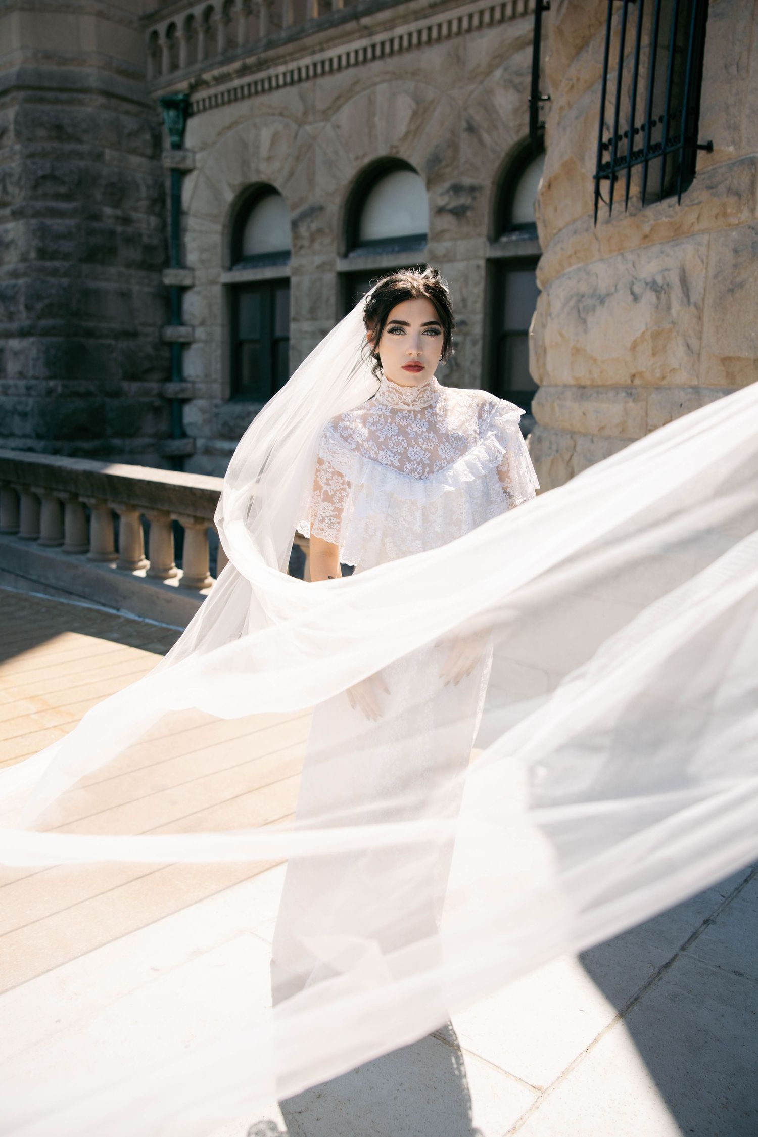 Bride in high-neck lace gown with dramatic flowing veil poses near ornate stone building exterior.