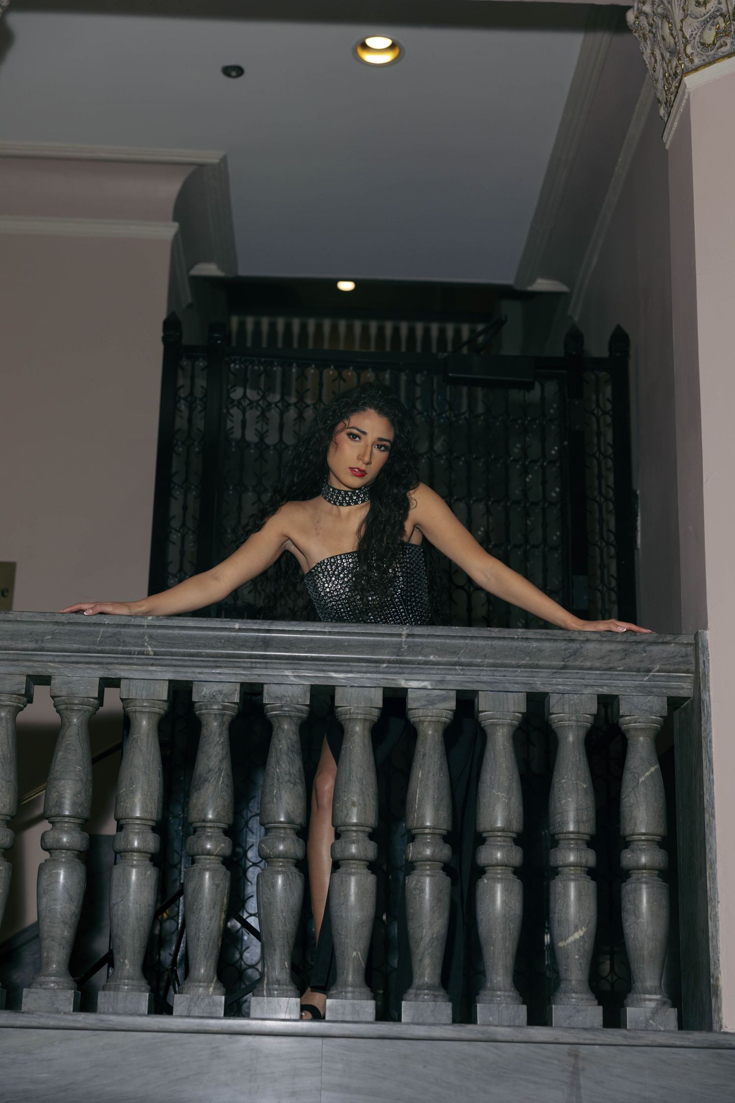 Woman in black sequin dress poses on a marble balcony railing at night.
