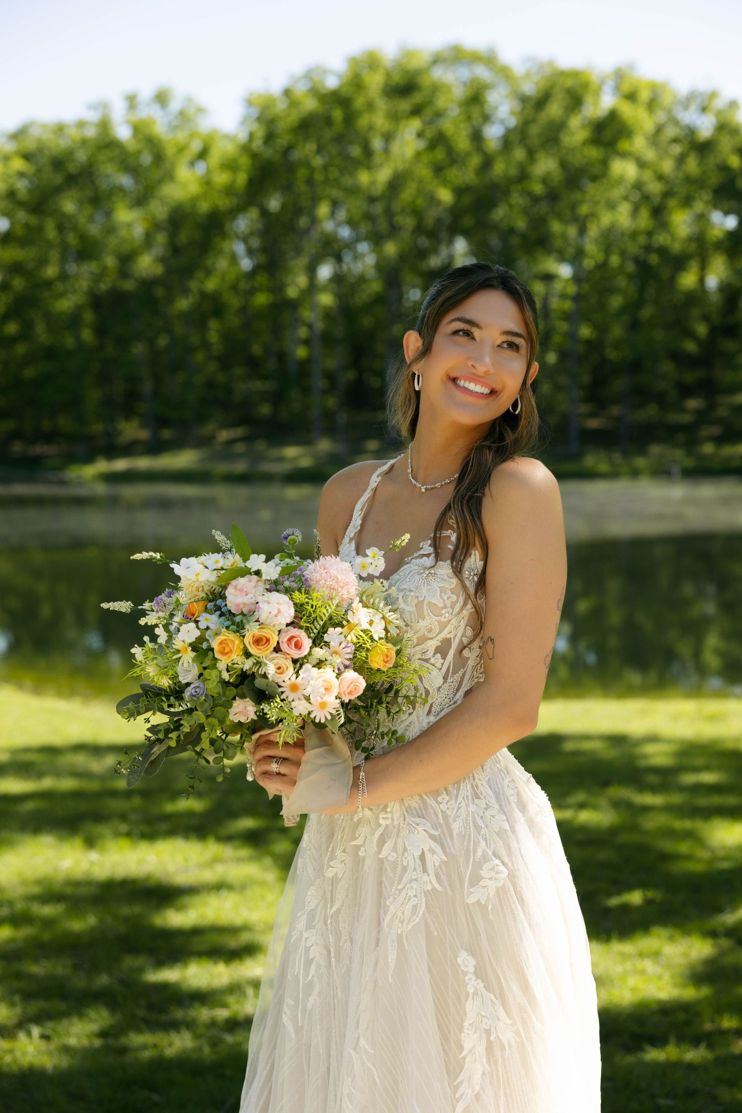 Smiling bride in white lace gown holds colorful bouquet outdoors near pond with lush green trees.