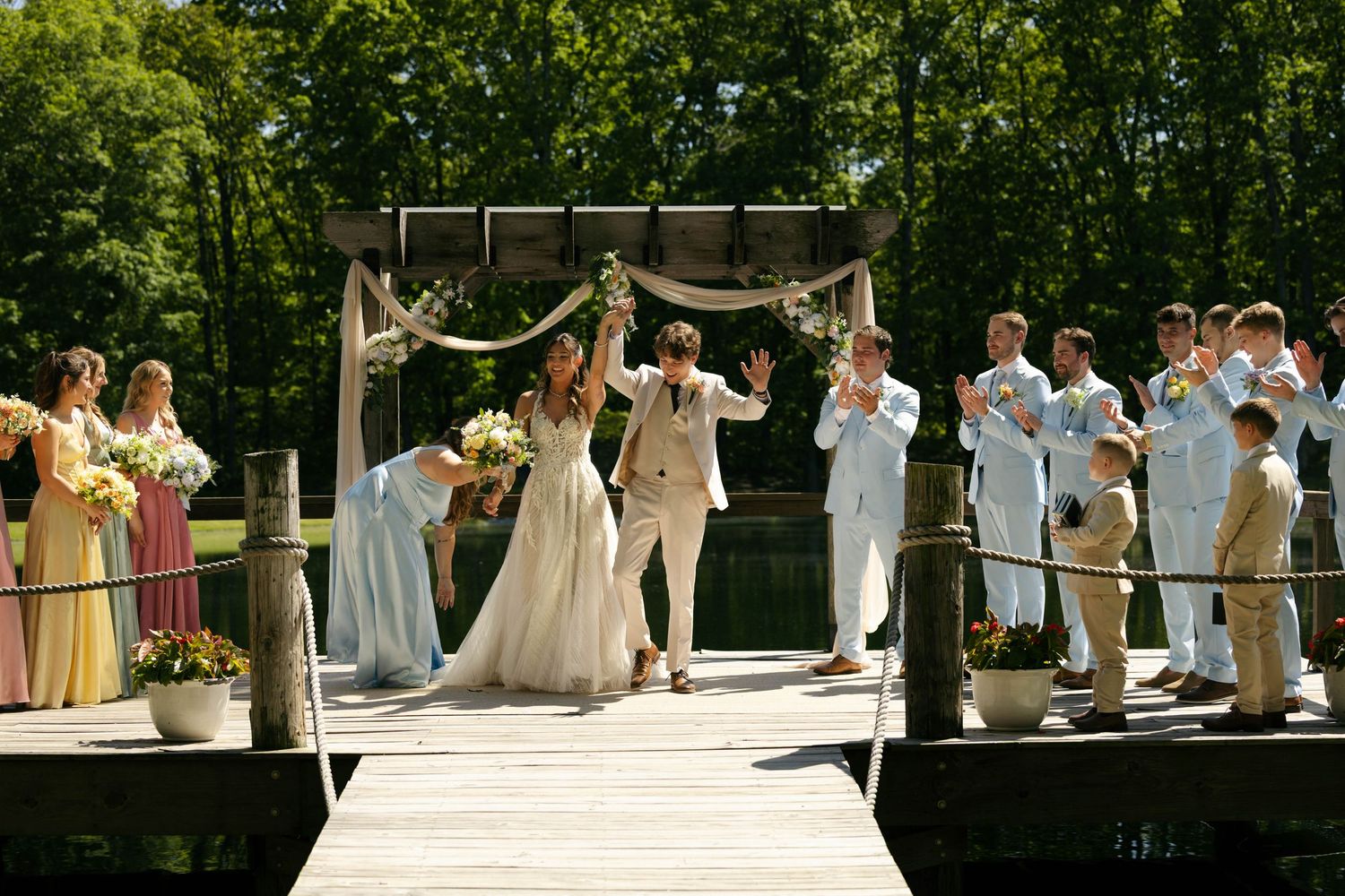 Outdoor wedding ceremony on a wooden dock with bride and groom celebrating, surrounded by wedding party and lush green trees.