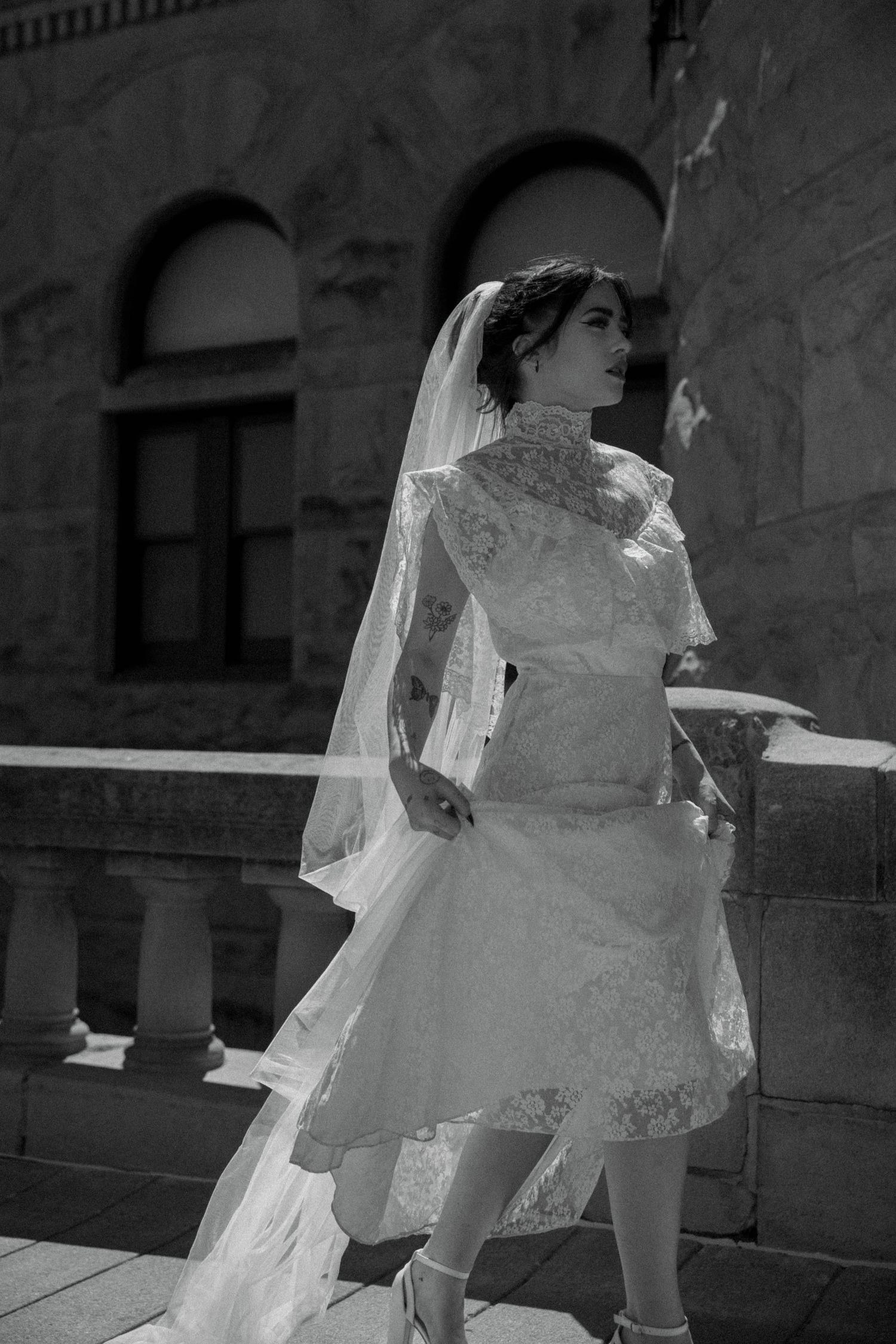 Black and white photo of tattooed bride in lace midi dress and veil on stone balcony.