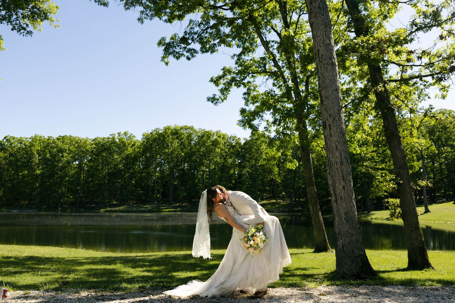 Bride in white wedding gown and veil bowing gracefully near a serene lake surrounded by lush green trees on a sunny day.