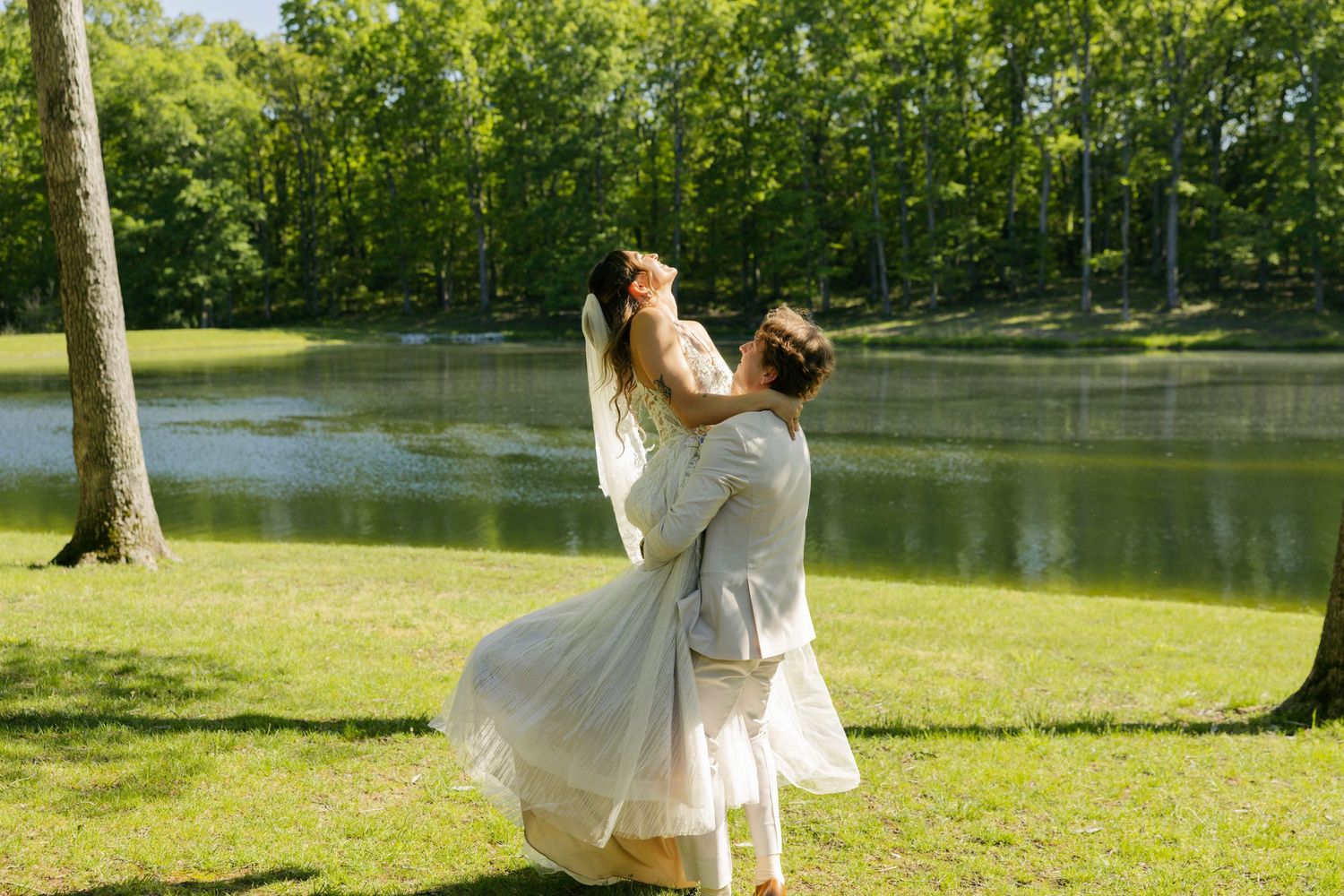 Couple in white wedding attire dancing and spinning joyfully by a serene lake surrounded by lush green trees.