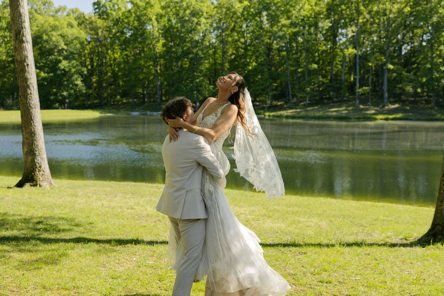 Bride and groom sharing a romantic embrace in a lush green park by a serene pond on their wedding day.