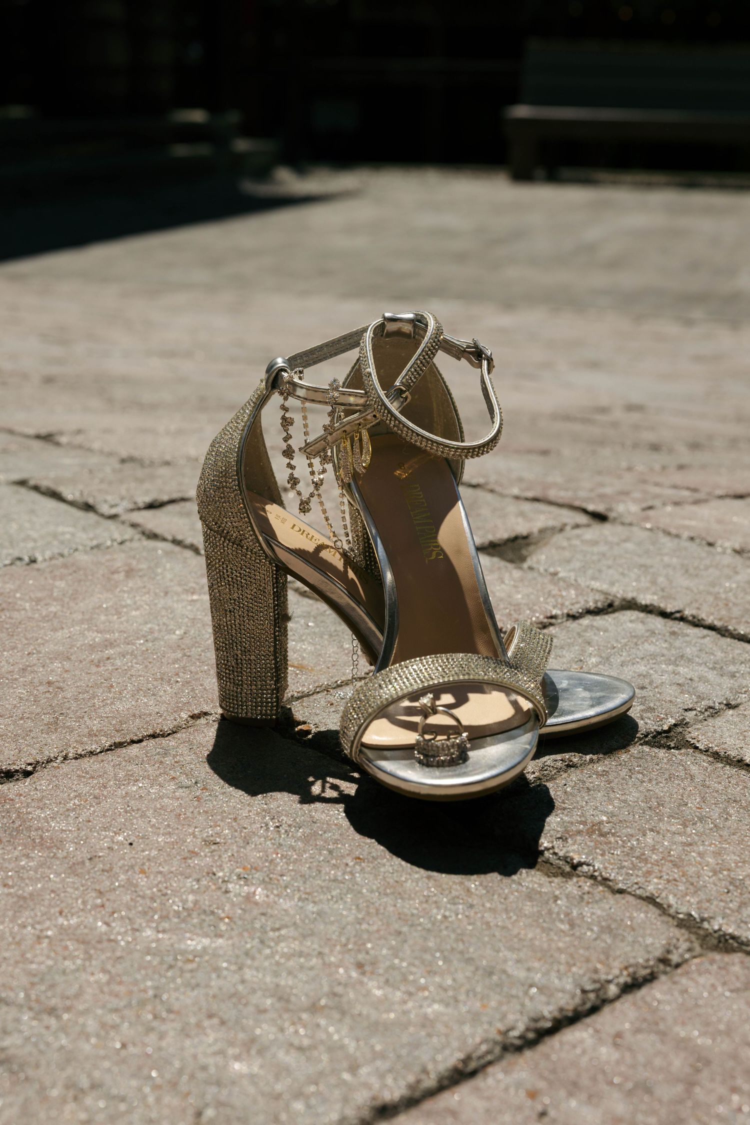Gold glittery block heel sandals with ankle strap and chain detail on stone pavement in sunlight.