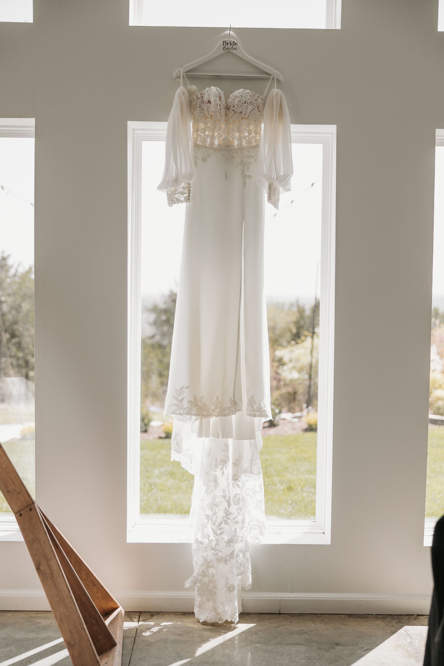 Elegant white wedding gown with lace details and sheer sleeves hanging in sunlit window.