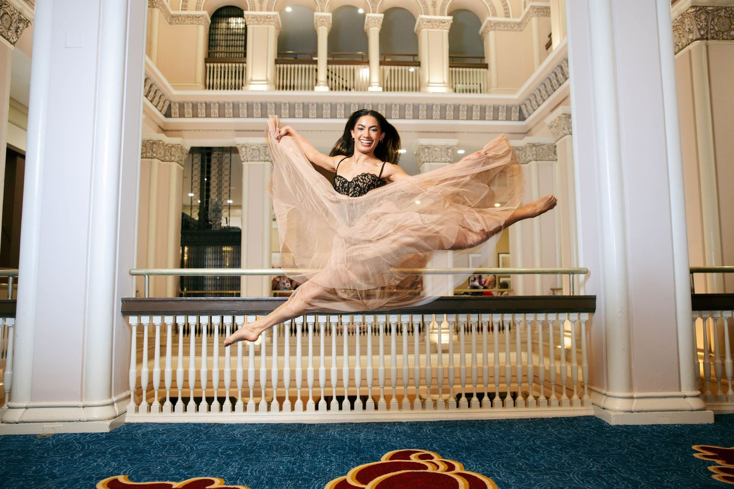 A ballet dancer leaps gracefully in a flowing beige gown inside an elegant grand hall with white columns.